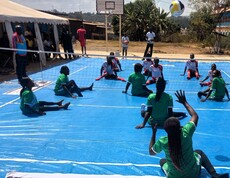 Frauenmannschaften spielen Sitzvolleyball auf einem Spielfeld im Freien, während Zuschauer*innen und Schiedsrichter das Spiel verfolgen.