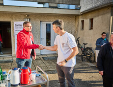 Zwei Männer schütteln sich die Hände vor einem Gebäude, in dem Caritas Soforthilfe organisiert. Einer trägt eine rote Caritas-Jacke, während der andere ein weißes T-Shirt anhat. Im Hintergrund sind weitere Personen und ein Caritas-Banner sichtbar.