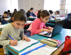 In einem Klassenzimmer sitzen zwei Schülerinnen an einem Tisch und machen Schulaufgaben. Eine Schülerin arbeitet an einem Laptop.