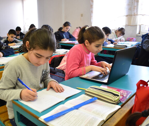 In einem Klassenzimmer sitzen zwei Schülerinnen an einem Tisch und machen Schulaufgaben. Eine Schülerin arbeitet an einem Laptop.