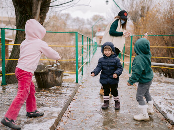 Drei Kinder, die Regenkleidung tragen, spielen vor einem Zentrum im Regen mit ihrer Betreuerin.