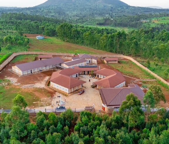  Drohnenaufnahme des vollständigen Gebäudekomplexes des Institut Supérieur de Nutrition et Paramédical de Simba inmitten grüner Hügel und Waldlandschaft; mehrere Backsteingebäude mit roten Dächern und Solarpanelen sind um einen zentralen Innenhof gruppiert, umgeben von einer Umfassungsmauer und terrassierten Feldern.