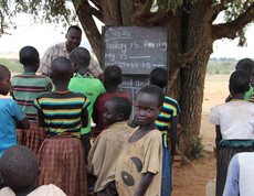 Eine Gruppe von Kindern sitzt unter einem Baum vor einer Tafel, während ein Lehrer im Freien unterrichtet.