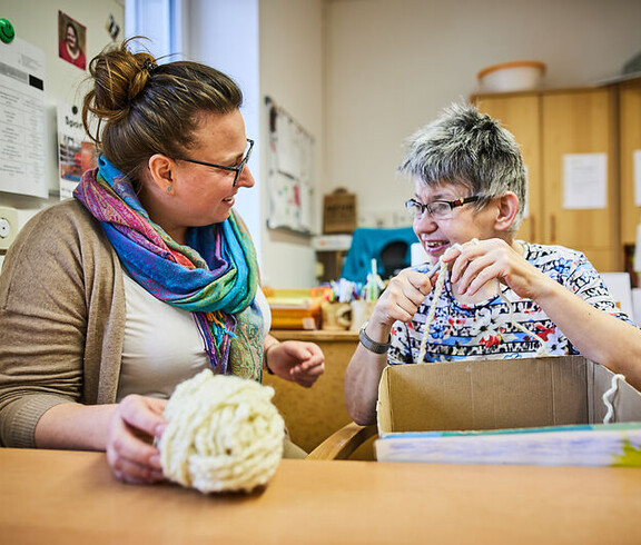 Zwei Frauen arbeiten gemeinsam an einem Tisch, eine hält Material in der Hand, beide lächeln