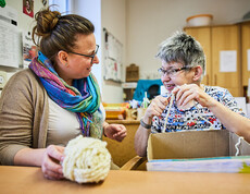 Zwei Frauen arbeiten gemeinsam an einem Tisch, eine hält Material in der Hand, beide lächeln