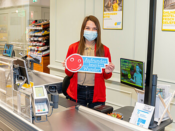 Eine Frau steht hinter der Supermarkt-Kassa, sie hält ein Schild in der Hand mit dem Schriftzug "Aufrunder bewirken Wunder".