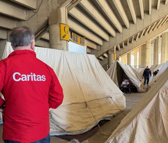 Zelte, die als Notunterkünfte dienen. Im Vordergrund ist ein Mann von hinten zu sehen, er trägt eine rote Jacke mit Aufschrift "Caritas".
