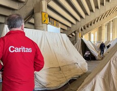 Zelte, die als Notunterkünfte dienen. Im Vordergrund ist ein Mann von hinten zu sehen, er trägt eine rote Jacke mit Aufschrift "Caritas".