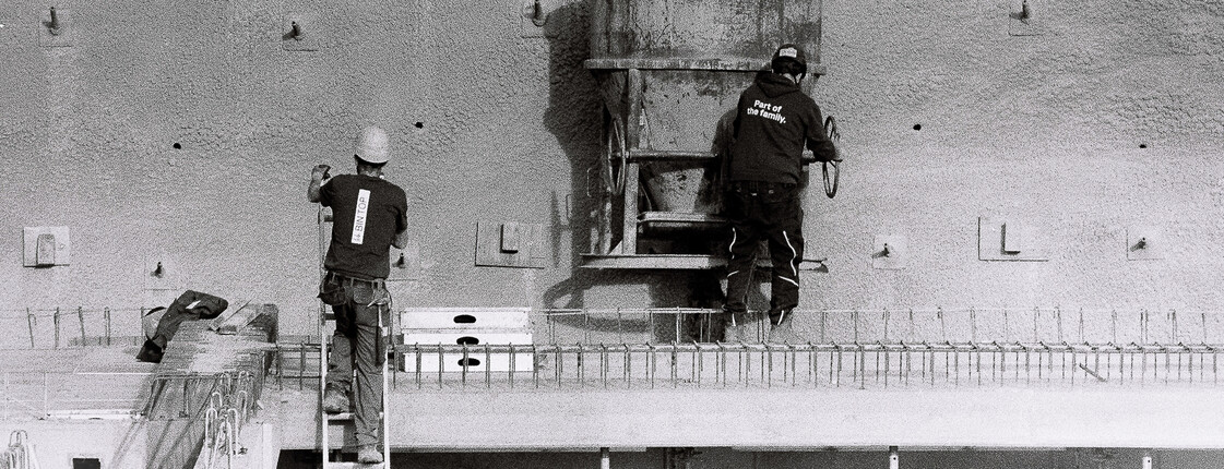 Zwei Bauarbeiter*in arbeiten an Betonwand mit Kran und Schalungselementen