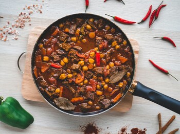 Integrationshaus Mittagstisch: Tischlein deck dich Obersicht einer Pfanne mit Chilli con Carne, die auf einem kleinen Holzbrette auf einem Esstisch steht. Drumherum liegen Zutaten wie Chillischoten und eine grüne Paprika.
