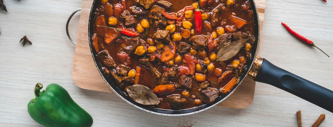 Obersicht einer Pfanne mit Chilli con Carne, die auf einem kleinen Holzbrette auf einem Esstisch steht. Drumherum liegen Zutaten wie Chillischoten und eine grüne Paprika.