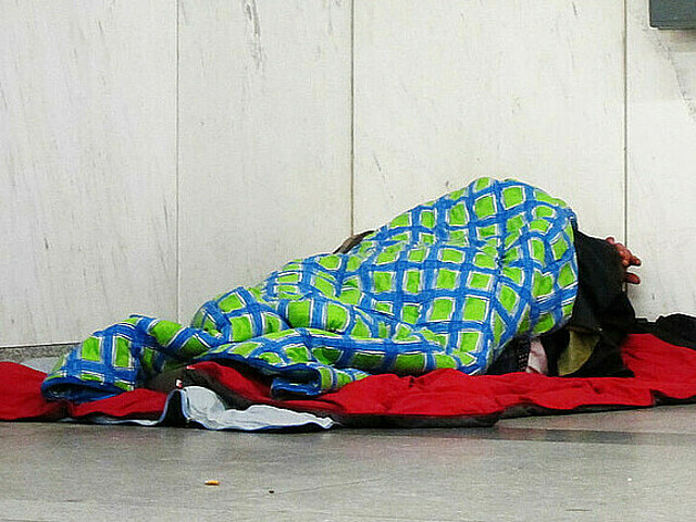 Man erkennt eine Person, dich zugedeckt mit Decken, sie liegt auf einem kalten Steinboden in der Ecke einer U-Bahn-Station