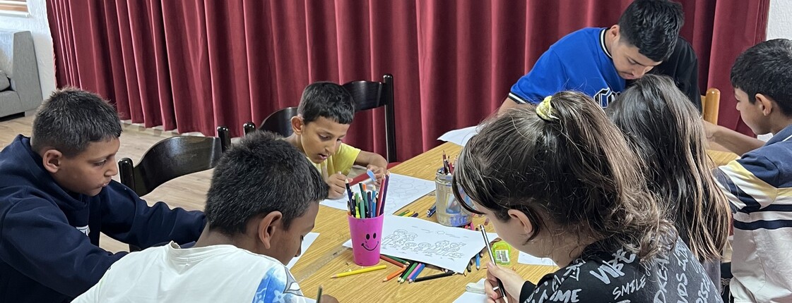 Mehrere Kinder sitzen um einen Tisch und malen mit Buntstiften auf Papier; auf dem Tisch stehen Becher mit Stiften.