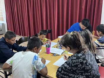 Mehrere Kinder sitzen um einen Tisch und malen mit Buntstiften auf Papier; auf dem Tisch stehen Becher mit Stiften.