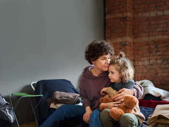 Eine Frau sitzt auf einer Bank, auf ihrem Schoß ein kleines Kind mit Kuscheltier in der Hand.