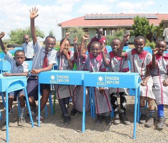 Eine Gruppe fröhlicher Kinder in Schuluniform jubelt und winkt in die Kamera, sitzend an neuen blauen Schulbänken mit der Aufschrift PACIDA im Schulhof der Tiigo Schule in Marsabit, Kenia; im Hintergrund ein Schulgebäude mit Solarpanelen.