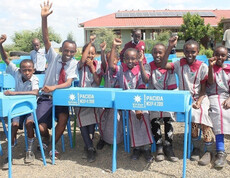 Eine Gruppe fröhlicher Kinder in Schuluniform jubelt und winkt in die Kamera, sitzend an neuen blauen Schulbänken mit der Aufschrift PACIDA im Schulhof der Tiigo Schule in Marsabit, Kenia; im Hintergrund ein Schulgebäude mit Solarpanelen.