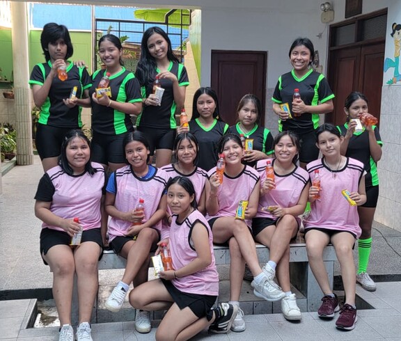 Zwei Gruppen von Mädchen und jungen Frauen in schwarzen und rosafarbenen Sporttrikots stehen lächelnd mit Getränken im Innenhof des Heims El Huambrillo für ein Gruppenfoto zusammen.