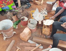 Mehrere teilweise zusammengebaute Metallkochöfen aus Zink- und Tonmaterialien stehen auf rotem Lehmboden; eine Person hält einen runden Metalldeckel in der Hand – Fertigung energieeffizienter Kochöfen im Rahmen des Caritas-Projekts in Burundi.