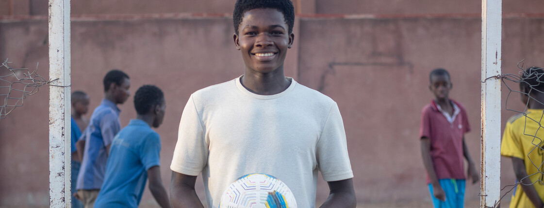 Portrait eines Jugendlichen, der ein weißes T-Shirt trägt und einen Fußball in den Händen hält. Im Hintergrund ist ein Fußballfeld auf dem Kinder Fußball spielen.