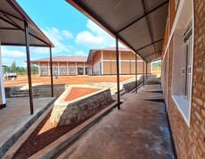  Blick über den gepflasterten Innenhof des neu errichteten Ausbildungszentrums in Simba: überdachte Verbindungsgänge aus Metall, Backsteingebäude mit roten Wellblechdächern und Natursteinmauern unter blauem Himmel – Institut Supérieur de Nutrition et Paramédical de Simba, Burundi.