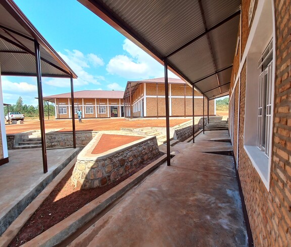  Blick über den gepflasterten Innenhof des neu errichteten Ausbildungszentrums in Simba: überdachte Verbindungsgänge aus Metall, Backsteingebäude mit roten Wellblechdächern und Natursteinmauern unter blauem Himmel – Institut Supérieur de Nutrition et Paramédical de Simba, Burundi.