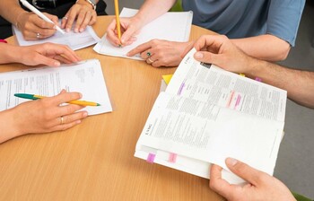 Man sieht die Hände von vier Personen, die an einem Tisch sitzen und Stifte, Zettel und Bücher halten. Die Gesichter sind nicht sichtbar. 