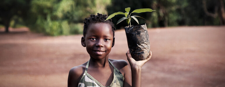 Kind hält Setzling in schwarzem Pflanzsack und lächelt. Hintergrund: rote Erde, Bäume, Natur.