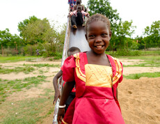 Ein lachendes Mädchen im roten Kleid steht vor einer Rutsche, auf der weitere Kinder sitzen, auf dem Spielplatz der Gumbo Primary School in Südsudan.