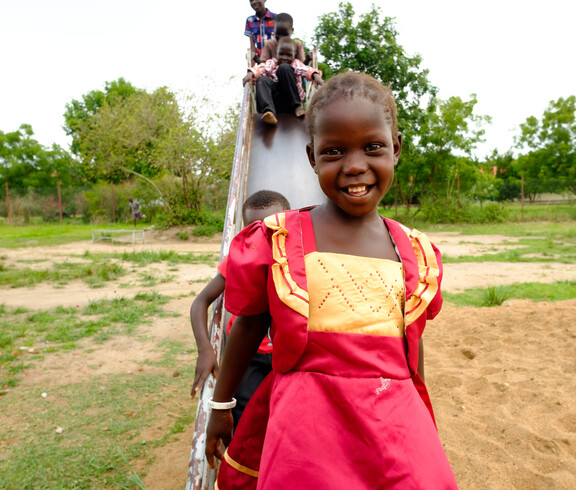 Ein lachendes Mädchen im roten Kleid steht vor einer Rutsche, auf der weitere Kinder sitzen, auf dem Spielplatz der Gumbo Primary School in Südsudan.