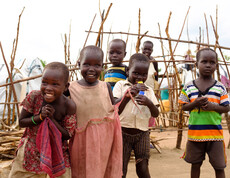 Gruppe lachender Kleinkinder vor einem Holzzaun und provisorischen Zelten im Camp Gumbo, Südsudan.