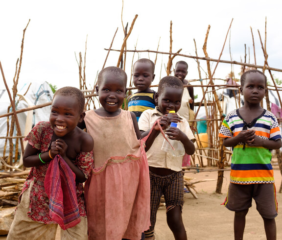 Gruppe lachender Kleinkinder vor einem Holzzaun und provisorischen Zelten im Camp Gumbo, Südsudan.