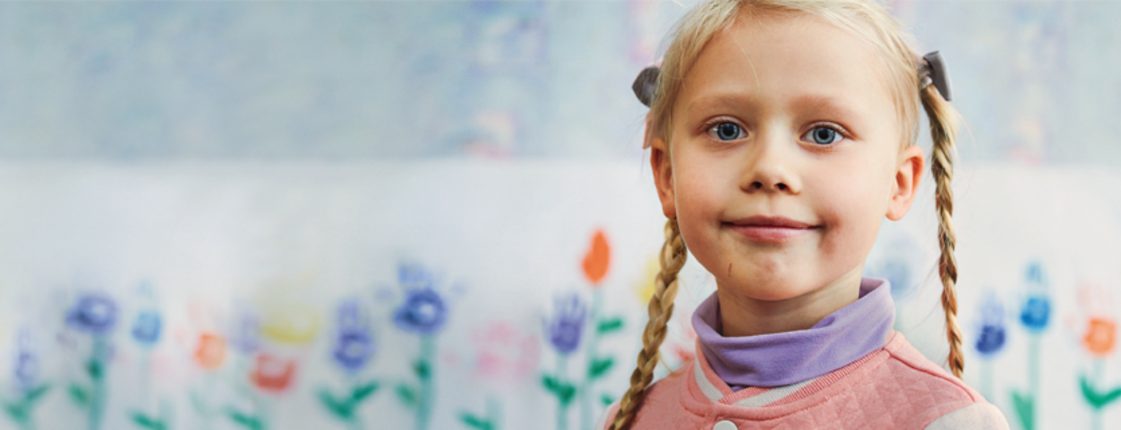 Patenschaften für Kinder weltweit Ein junges Mädchen mit blonden Zöpfen steht vor einer bunten Wand mit gemalten Blumen und lächelt freundlich.