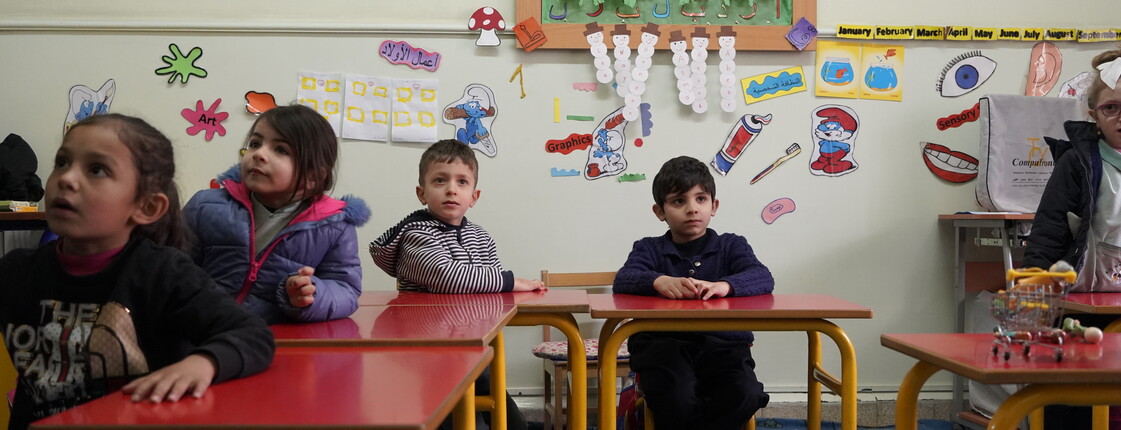 Vier Kinder sitzen aufmerksam an roten Tischen in einem bunt dekorierten Klassenraum der Caritas