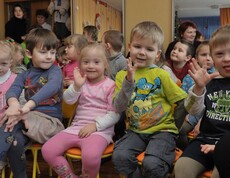 Kinder sitzen gemeinsam im Kindergarten und winken, darunter auch Kinder mit Behinderungen