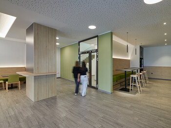 Innenansicht Aula Caritas Schule Wiener Neustadt, 2 Mädchen gehen zur Glastür Richtung Stiegenhaus