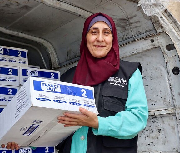 Eine Mitarbeiterin der Caritas Jerusalem steht vor einem Lieferwagen und hält eine Kiste mit Babynahrung in den Händen. Im Fahrzeug sind gestapelte Hilfspakete zu sehen.