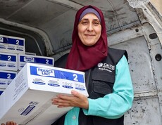 Eine Mitarbeiterin der Caritas Jerusalem steht vor einem Lieferwagen und hält eine Kiste mit Babynahrung in den Händen. Im Fahrzeug sind gestapelte Hilfspakete zu sehen.