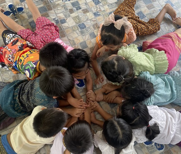 Mehrere Kinder liegen im Kreis auf dem Boden und halten ihre Hände in der Mitte zusammen.