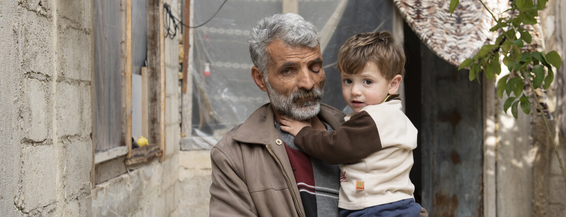 Älterer Mann mit kleinem Kind vor einem beschädigten Haus in Syrien.