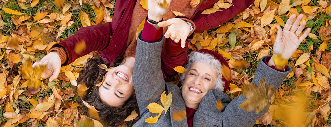 Eine ältere und eine jüngere Frau liegen auf dem Rücken am Boden und werfen buntes Herbstlaub in die Luft.