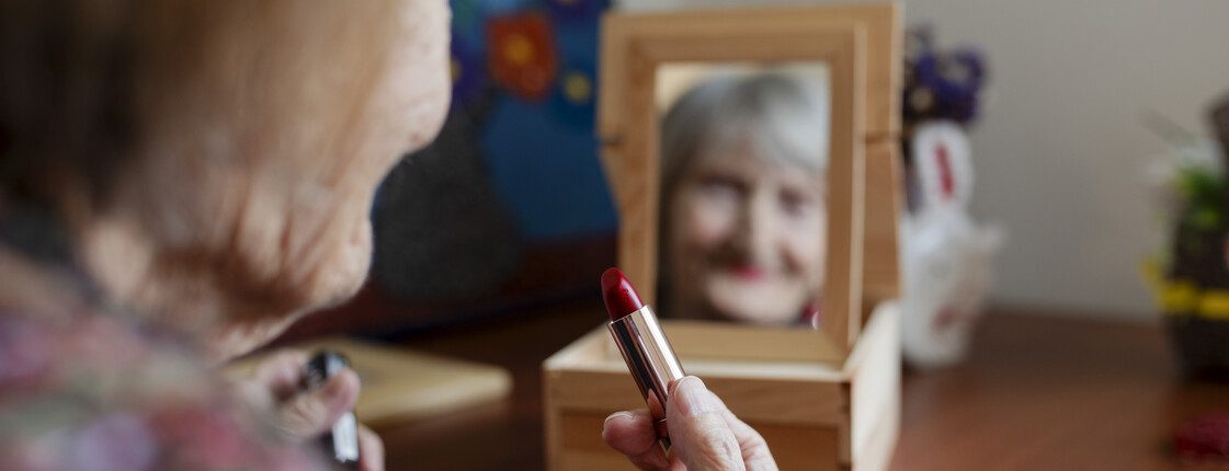Eine alte Dame sitzt vor einem kleinen Spiegel und schminkt sich. Wir sehen sie von hinten, jedoch spiegelt sich ihr Gesicht im kleinen Spiegel. Sie hat einen roten Lippenstift in der Hand. 
