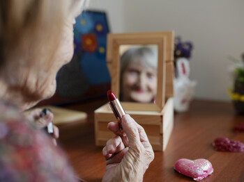 Eine alte Dame sitzt vor einem kleinen Spiegel und schminkt sich. Wir sehen sie von hinten, jedoch spiegelt sich ihr Gesicht im kleinen Spiegel. Sie hat einen roten Lippenstift in der Hand. 