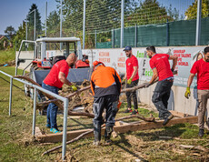Freiwillige Helfer räumen Baumstämme und Schutt auf einem Sportplatz in Hofstetten-Grünau. Sie tragen rote Caritas-T-Shirts, und im Hintergrund steht ein Traktor bereit, um die Überreste abzutransportieren. Die Helfer arbeiten gemeinsam daran, die Hochwasserschäden zu beseitigen.