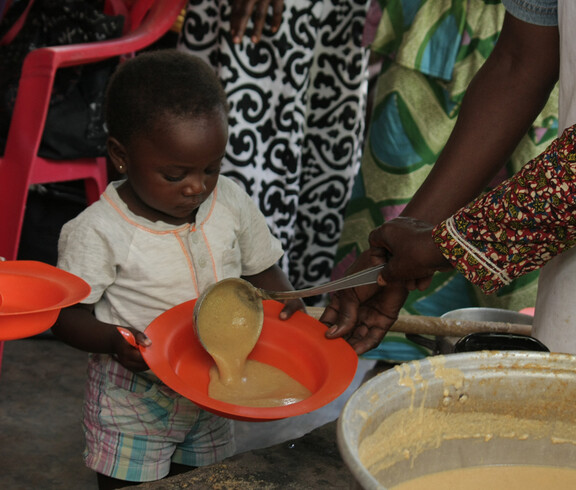 Ein kleines Kind hält einen orangefarbenen Teller, während ihm in der Ernährungsstation nahrhafter Brei aus einem großen Topf serviert wird