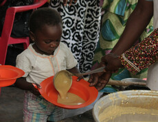 Ein kleines Kind hält einen orangefarbenen Teller, während ihm in der Ernährungsstation nahrhafter Brei aus einem großen Topf serviert wird