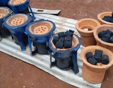 Mehrere fertig montierte energieeffiziente Kochöfen aus blauem Metall und Ton stehen nebeneinander; in einem Ofen liegen schwarze Kohlebriketts aus Pflanzenabfällen, daneben weitere Tonöfen ohne Metallmantel.