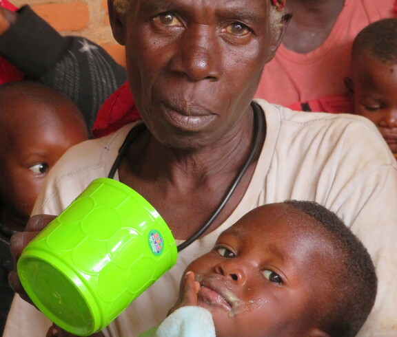 Nahaufnahme eines kleinen Kindes, dem aus einem grünen Plastikbecher Nahrungsbrei gereicht wird; das Kind trinkt konzentriert, ein Waschlappen wischt den Mund – Versorgung im Ernährungszentrum Gitega, Burundi.