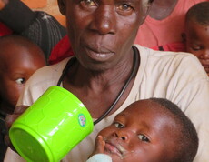 Nahaufnahme eines kleinen Kindes, dem aus einem grünen Plastikbecher Nahrungsbrei gereicht wird; das Kind trinkt konzentriert, ein Waschlappen wischt den Mund – Versorgung im Ernährungszentrum Gitega, Burundi.