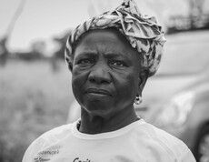 Eine Frau blickt in die Kamera; sie steht draußen und trägt ein Kopftuch sowie ein T-Shirt mit Projektaufdruck.